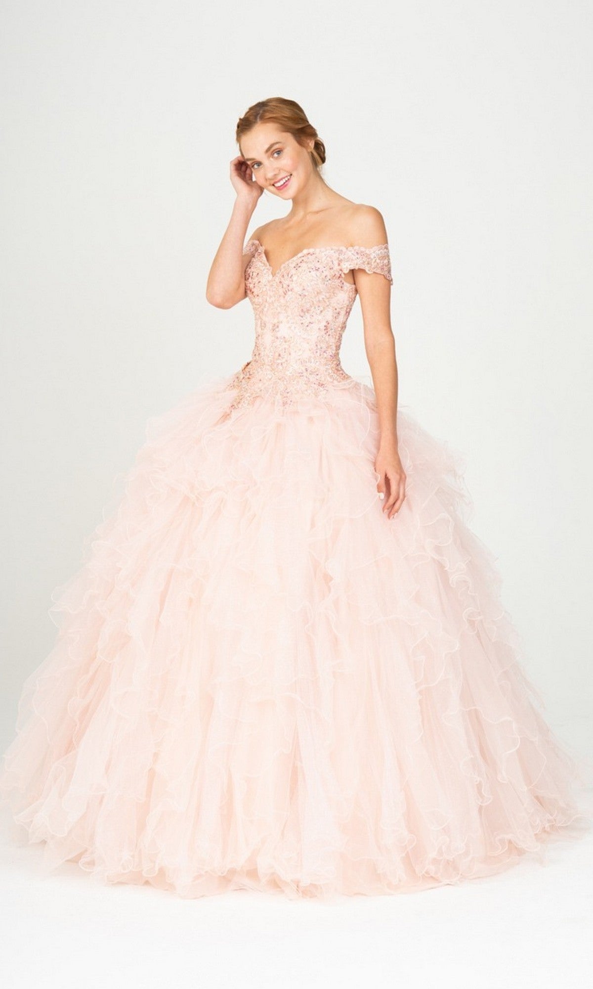 A woman poses against a white background in the Quinceanera Dress 9313 By Eureka, featuring a blush hue, beaded off-the-shoulder bodice, and a ruffled ball gown skirt.